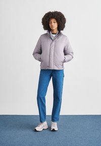 Χειμωνιάτικο μπουφάν σε ανοιχτό μωβ χρώμα με ψηλό γιακά, κούμπωμα με πιετάκια μπροστά και δύο πλαϊνές τσέπες. Φορεμένο με μπλε τζιν παντελόνι και γκρι αθλητικά παπούτσια.