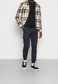 Randig flanellskjorta i krämfärg, svart och orange; svart T-shirt; marinblå cargobyxor; svarta sneakers med vita sulor och vita strumpor.