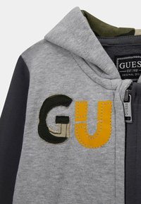 Haut à capuche zippé gris avec des accents noirs et olive. Présente le « GU » brodé en vert et jaune. Comprend une étiquette en tissu avec la marque « Guess ». 