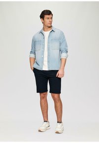 Chaqueta de mezclilla azul clara sobre una camiseta blanca, combinada con pantalones cortos azul marino oscuros y zapatillas blancas. La chaqueta cuenta con dos bolsillos en el pecho.