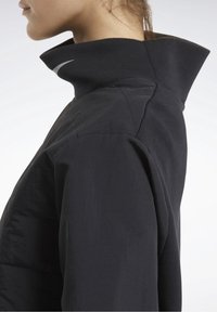 Chaqueta negra con un alto cuello, fabricada con un tejido suave y con acentos texturizados en los hombros. Diseño simple con detalles mínimos.
