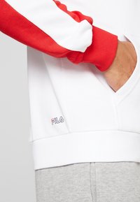 Sudadera blanca con mangas rojas, que presenta un puño acanalado. Logotipo bordado en azul y rojo en el frontal. Tela suave con detalle de bolsillo.