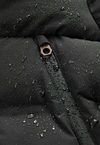 Nahaufnahme von Wassertropfen auf einem schwarzen wasserdichten Jackenstoff mit sichtbarem Reißverschluss und Metallziehhilfe.