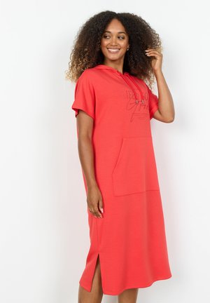 Frau mit lockigem Haar, die ein knielanges korallenfarbenes Kapuzenkleid mit kurzen Ärmeln und einer Fronttasche trägt, lächelnd und ihr Haar berührend.