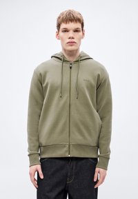 Felpa con cappuccio verde oliva con zip, cappuccio regolabile con coulisse, polsini e orlo a costine. Presenta un logo sul petto a sinistra e una texture di tessuto liscia.