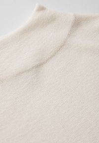 Tessuto color crema con una finitura morbida e texturizzata e un collo arrotondato. Il materiale sembra leggero e leggermente elasticizzato.