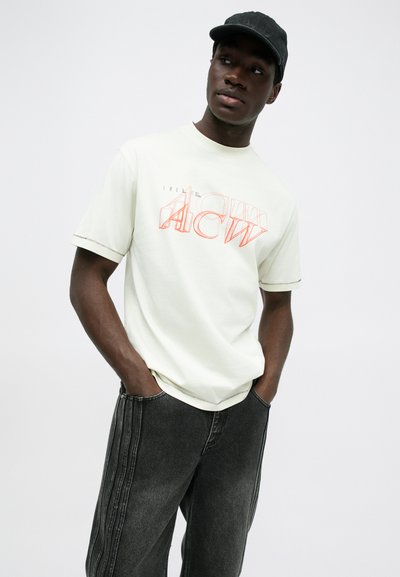 Camiseta de color claro y manga corta con texto gráfico en naranja. Llevada con jeans de mezclilla oscuros que presentan franjas laterales y costuras visibles.