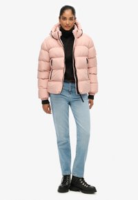 Roze gewatteerde jas met een capuchon, ritssluiting aan de voorkant en zijzakken, gedragen over een zwarte coltrui, gecombineerd met lichtblauwe jeans en zwarte laarzen.
