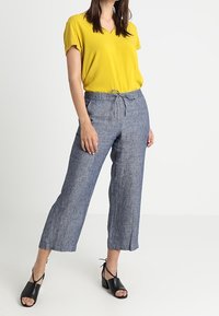Blusa gialla a scollo a V con maniche corte abbinata a pantaloni in lino blu a vita media e a gamba larga, accessorata con sandali neri con tacco.