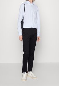 Hellblaues Hemd mit langen Ärmeln, schwarze Hose und weiße Sneaker mit beigen Akzenten. Eine Umhängetasche fügt praktische Details hinzu.