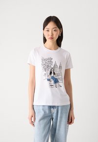 T-shirt en coton blanc avec un imprimé graphique d'une fille assise au milieu de bâtiments de la ville et d'arbres, col rond, manches courtes, coupe décontractée.