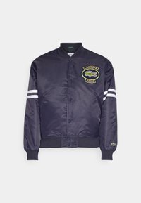 Námořnická saténová bomber bunda s zeleným logem krokodýla Lacoste, bílými pruhy na rukávech a žebrovanými manžetami. Zapínání na cvoky.