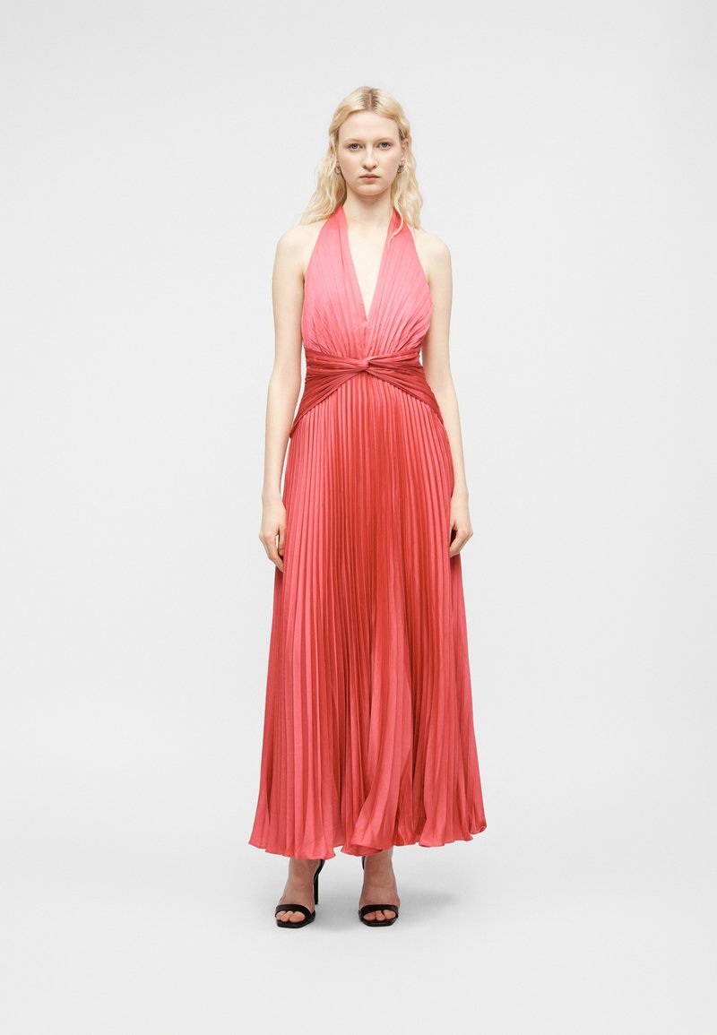 Blonde Frau steht in einem ärmellosen, plissierten, korallenfarbenen Kleid mit Neckholder und gedrehtem Taillendetail sowie schwarzen offenen Absatzschuhen vor weißem Hintergrund.