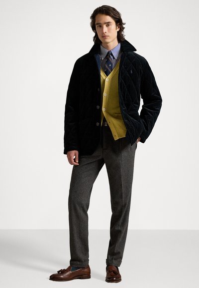 Melna izšūta jaka virs sinepju krāsas džemperis, kārtota ar svītrotu krekla un kaklasaiti. Pelēka svārku bikses un brūnā loafers piepilda apģērbu.