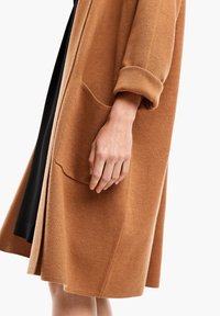 Hnědý kabát z vlněné směsi s nařasenými rukávy, velkou přední kapsou a měkkým, strukturovaným povrchem. Vnitřní vrstva je černá a ukazuje vrstvený design.