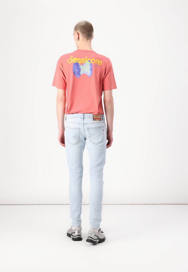 Rotes T-Shirt mit blauer Grafik "dieselcore" und hellblauen Skinny-Jeans. Die Jeans haben eine subtile, abgeriebene Textur und Details an der Gesäßtasche.