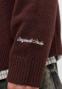 Burgundska pletena jopica z rebrastimi manšeti, ki ima na rokavu vezen napis "Originals Studio" v beli barvi. Mehka tekstura.
