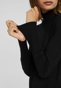 Zwarte coltrui met geribbelde manchetten, nauwsluitende mouwen en een gladde textuur. Focus op de details rond de handen.