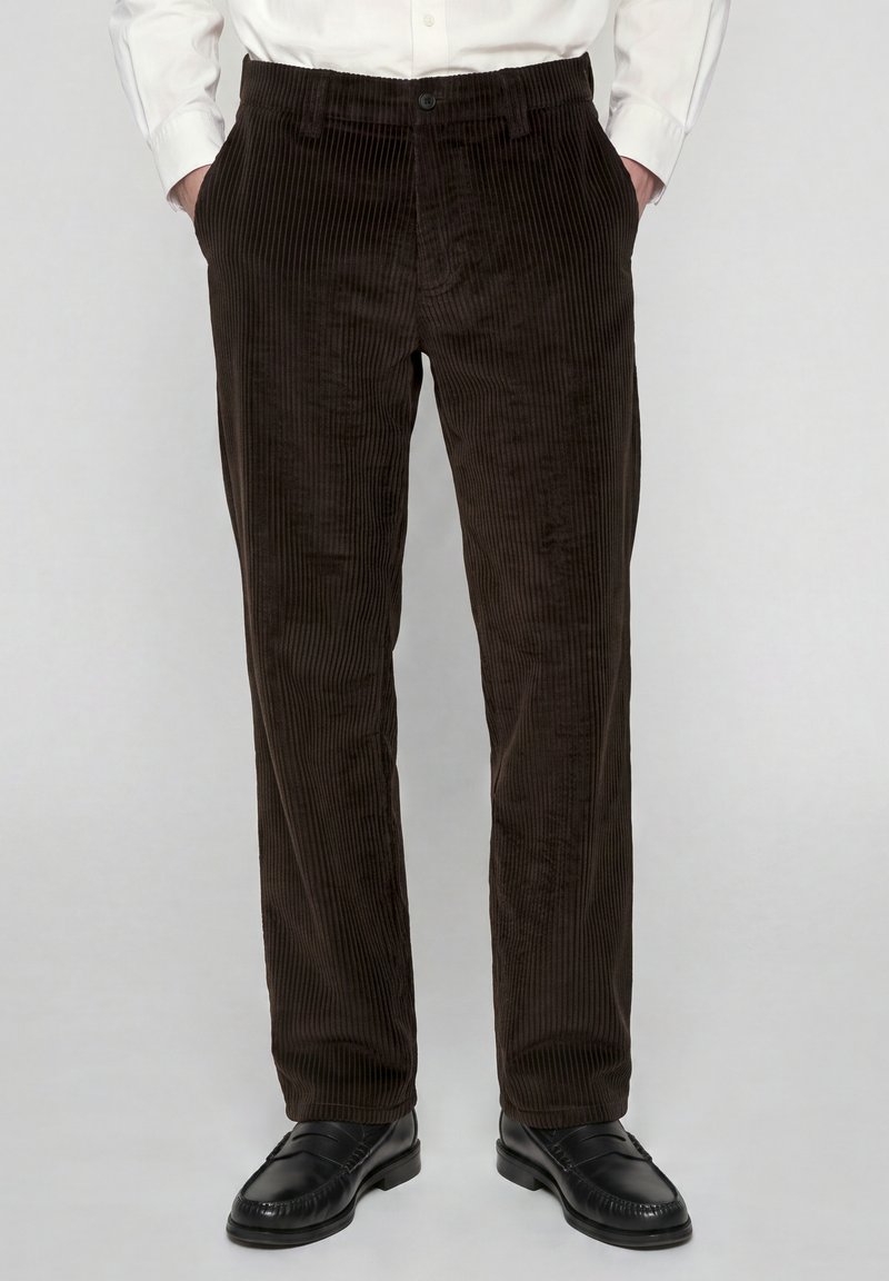 Un homme porte un pantalon en velours côtelé brun foncé, une chemise blanche rentrée, avec des mocassins en cuir noir, debout les mains dans les poches.
