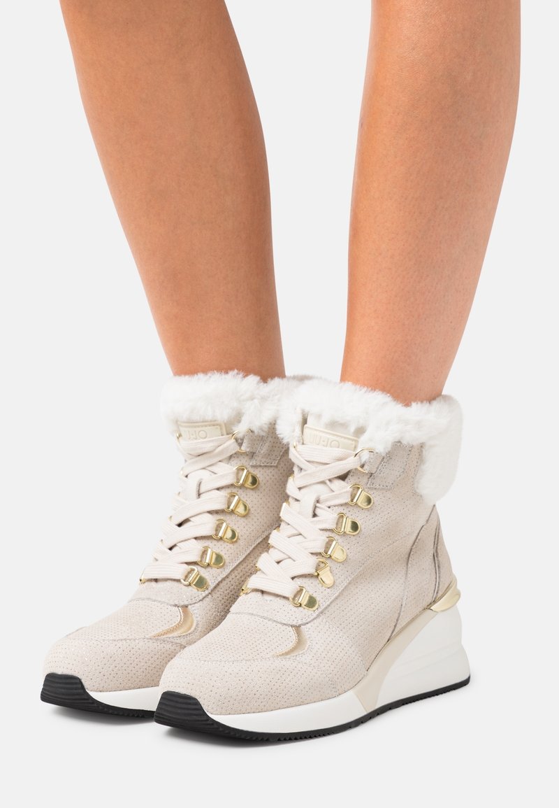 Chaussures montantes beiges avec une finition texturée, des accessoires en or et un col en fausse fourrure blanche, dotées d'une semelle compensée et d'un design épuré.