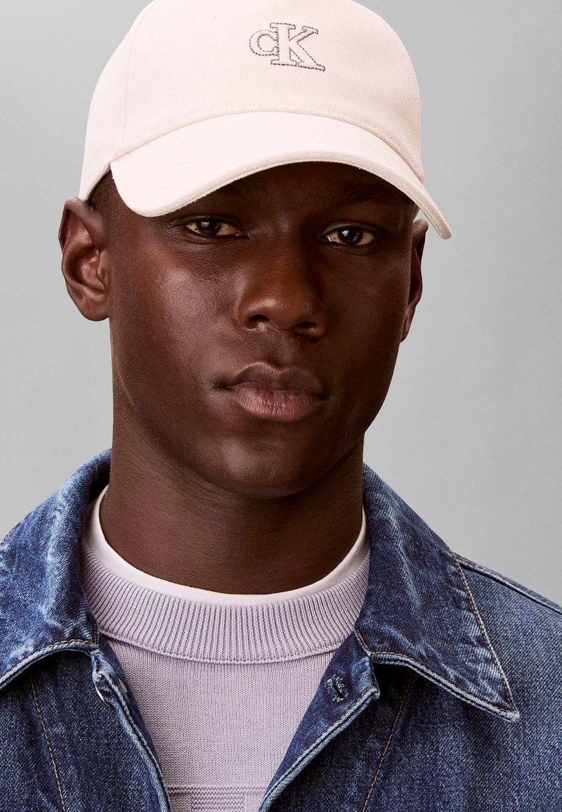 Jeune homme portant une casquette Calvin Klein beige clair, un pull col rond blanc et une veste en jean bleue sur fond gris uni.