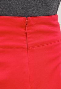 Tissu rouge texturé avec une finition lisse. Présente une fermeture éclair centrale à l'arrière et des coutures surpiquées pour un détail supplémentaire.