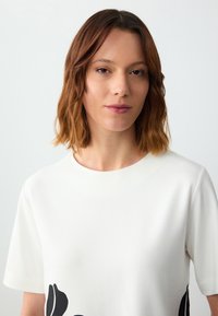 Weißes Kurzarmoberteil mit rundem Ausschnitt und einem schwarzen Blumenmuster auf der linken Seite. Glatte Stoffoberfläche mit leichtem Glanz.