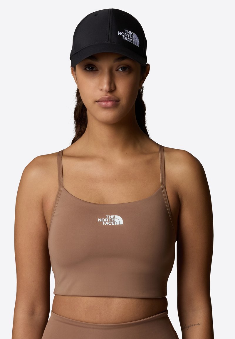 Top deportivo corto marrón con tirantes finos que presenta un logo blanco. El modelo lleva una gorra negra con el mismo logo. Textura suave.
