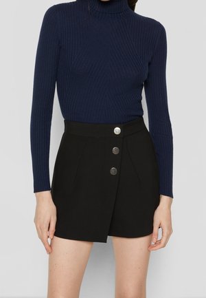 Vrouw draagt een marineblauwe geribbelde coltrui en zwarte shorts met hoge taille en drie zilveren knopen aan de voorkant.