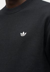Schwarzer Sweatshirt mit kleinem weißem Adidas-Trefoil-Logo, das auf der linken Brust eingestickt ist.