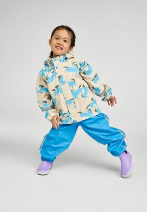 Laps naeratab ning teeb mängulise poosi, kandes beežikat jope siniste loomamustritega, erk siniseid pükse ja lilla kummisaapaid.