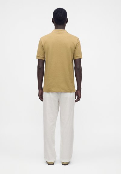 Homme à la peau foncée portant un polo à manches courtes couleur moutarde et un pantalon blanc, debout dos à un fond blanc uni.
