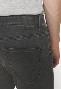 Detailní záběr na osobu v tmavě šedých jeansách s viditelnou zadní kapsou a černou nášivkou s nápisem "Only & Sons" na pase.
