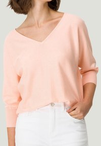 Femme portant un pull V rose clair à manches trois-quarts rentré dans un pantalon blanc taille haute, main dans la poche.