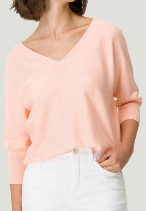 Femme portant un pull V rose clair à manches trois-quarts rentré dans un pantalon blanc taille haute, main dans la poche.