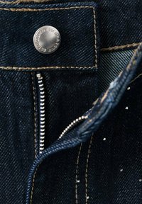 Jeans de mezclilla azul oscuro que cuentan con un botón y cremallera de metal plateado, con costuras marrón claro y pequeños detalles metálicos cerca de la cremallera.