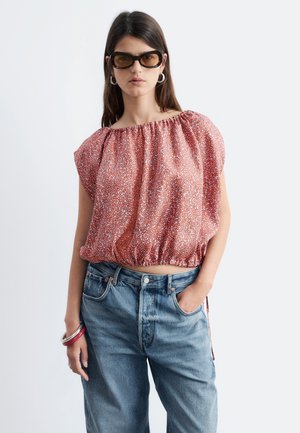 Frau trägt große Sonnenbrille, eine rote gemusterte schulterfreie Bluse, blaue hoch taillierte Jeans und Armbänder, posiert mit einer Hand in der Tasche.