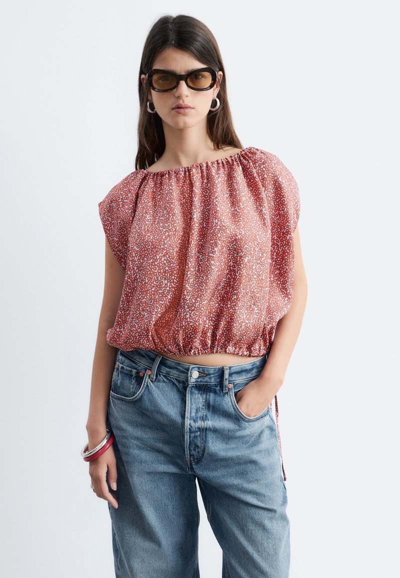 Femme portant de grandes lunettes de soleil, un chemisier rouge à motifs et épaules dénudées, un jean bleu taille haute et des bracelets, posant avec une main dans la poche.