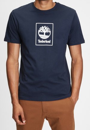 Man wearing a navy blue Timberland t-shirt with white tree logo and brown pants, hands relaxed at sides, against a plain background.