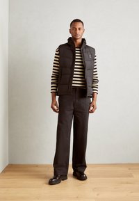 Black puffer vest over a striped black and white long-sleeve shirt, paired with dark trousers and black shoes, on a neutral background.
