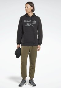 Sudadera de algodón negra con un logo de Reebok, combinada con pantalones de chándal verde oliva. Zapatillas grises con acentos negros y suela texturizada.