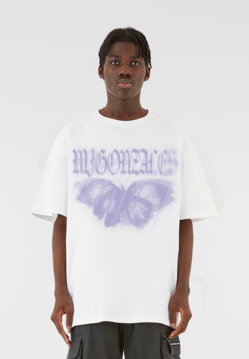 T-shirt bianco oversize con grafica di una farfalla viola e testo stilizzato. Materiale in cotone morbido con maniche corte e vestibilità rilassata.