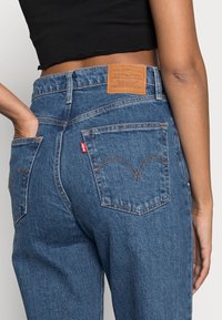 Jeans de mezclilla azul con cintura alta, que presentan un parche de cuero en la parte posterior, costuras naranjas y dos bolsillos traseros con costuras curvas.