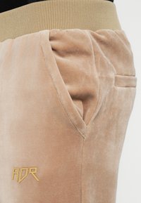 Joggers de terciopelo beige suave con una cinturilla acanalada en tono marrón, que cuentan con bolsillos laterales y un logo bordado en dorado en el muslo izquierdo.