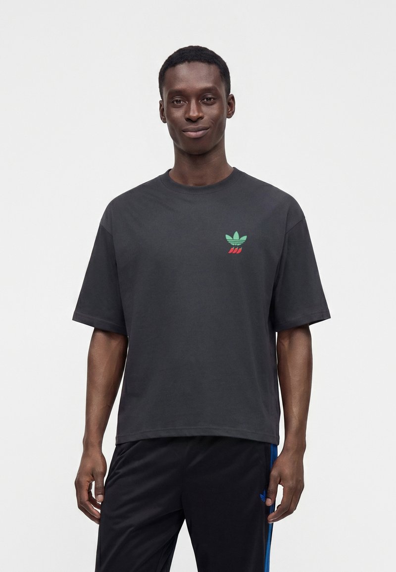 Man wearing a black oversized t-shirt with a small green and red logo on the left chest and black pants with blue stripes.