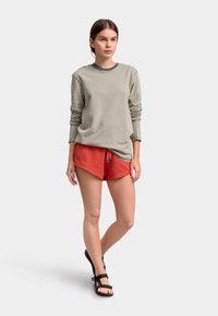 Felpa grigia chiara con maniche lunghe, abbinata a pantaloni corti rossi con coulisse. Il modello indossa sandali neri con due cinturini.