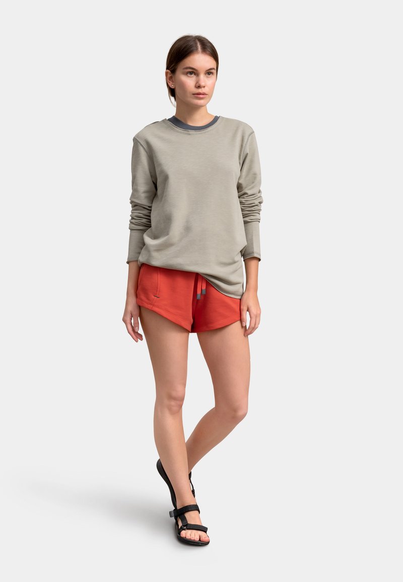 Felpa grigia chiara con maniche lunghe, abbinata a pantaloni corti rossi con coulisse. Il modello indossa sandali neri con due cinturini.