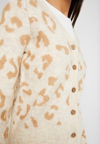 Cárdigan beige con una suave tela texturizada y un estampado de leopardo en marrón. Cinco botones de madera adornan el frente para el cierre.