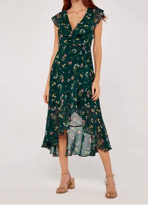Woman wearing a dark green floral wrap dress with ruffled sleeves and asymmetrical hem paired with beige block heel sandals.