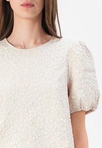 Béžový květinový vyšívaný top s krátkými nafouklými rukávy a kulatým výstřihem. Látka má texturované květinové vzory po celém povrchu.
